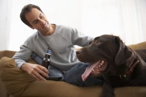 Man holding bottle relaxing on sofa with dog, smiling